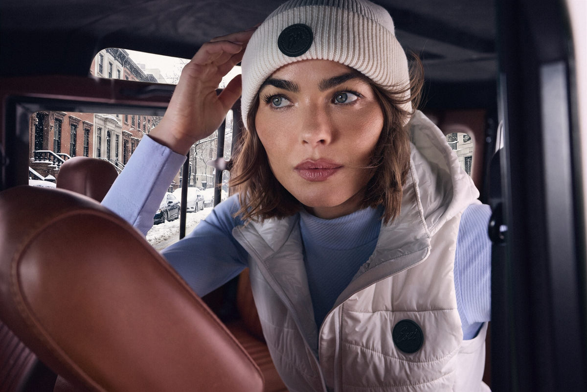 Woman in a Vanilla Teton Beanie and Aurora Vest, seen through the window of a vintage truck